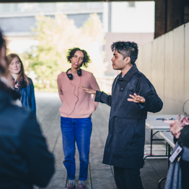 The artist in a black trenchcoat and pants outdoors speaking to an audience of playtesters