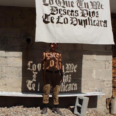 Sun shines through a canvas stencil rendering black letter text on a shirtless brown body—the text reads "Todo Lo Que Me Deseas Dios Te Lo Duplicara".