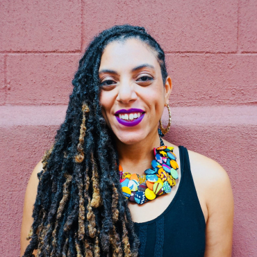 Locks pulled over to one side, multi-colored necklace made of West-African cloth buttons, black tank top, fuschia lipstick, and smiling golden brown face.