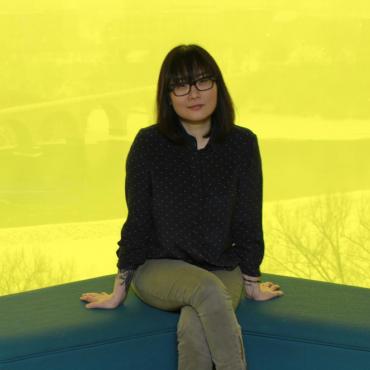 Su Hwang seated, top floor at the Guthrie Theater, bright yellow background overlooking the Stone Arch Bridge.