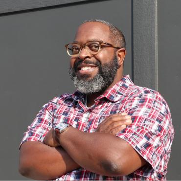 A Black man with black eyeglasses, short hair, and a fluffy black and gray beard is seen smiling with his arms crossed. He's wearing a red, white, and blue patterned shirt.