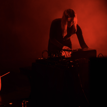 Leila Bordreuil, a 30 year old white woman with medium-long brown hair is facing the camera and playing electronic instruments on a black table. To her right, there is a cello on the floor. She is on a stage with red LED lighting and fog. The main colors of the photo are blacks and reds. 