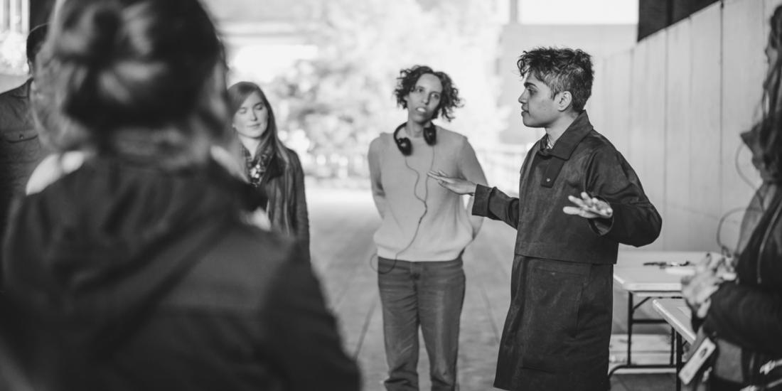 the artist in a black trenchcoat and pants outdoors speaking to an audience of playtesters