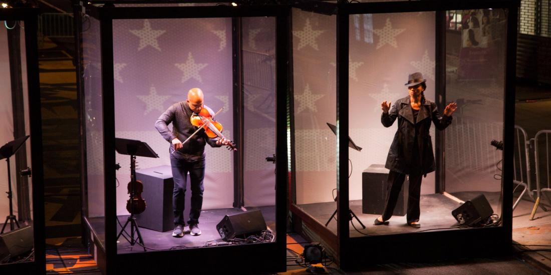 A violinist in a box next to a performer in a box on the street.