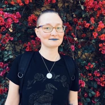 D. stands against a wall of vines with red flowers and dark green leaves. D. is wearing glasses, blue lipstick and a black t-shirt; they are smiling slightly.
