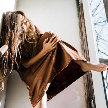 Leila is wearing a thobe - traditional Palestinian dress. This one is very old with faded red fabric and yellow embroidery. She is crouched in the corner of an outdoor staircase. A sliver of trees without leaves are visible. Her long brown hair covers her face. She pulls her knee towards her chest.