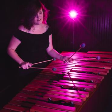 Jenny Klukken is playing the marimba with four mallets. She is wearing a black dress and a pink light is behind her.