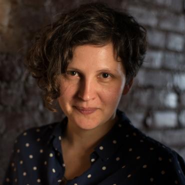 Anna Samo, a forty years old white woman with curly dark short hair, dressed into “used to be favourite” polka dot blue shirt, is smiling at the camera.