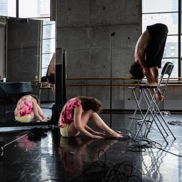 Two women are in the midst of a dance performance. One is standing on top of two high folding chairs. She is topless, wearing black pants, and her body is folded over her straight legs. The other woman is seated, hands and feet on the floor, with her torso and head folded over her legs. She is wearing yellow underwear and a pink tie-dye shirt. They are bowing to one another, both in profile, faces hidden. There is a mirror behind them reflecting a piano and the bodies. A microphone cord is tangled up.