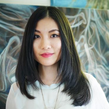 A young Asian woman with medium length black hair in a mint-colored shirt stands with her hands behind her back. In the background are paintings.