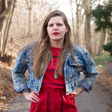 A white queer femme woman in red lipstick with long straight hair parted to the side in a red striped dress and short jean jacket stands with hands on hips on a path in a park. Brown trees without leaves, an evergreen, and grass are blurry in the background on the either side of the asphalt path. She has her hands with painted nails on her hips.