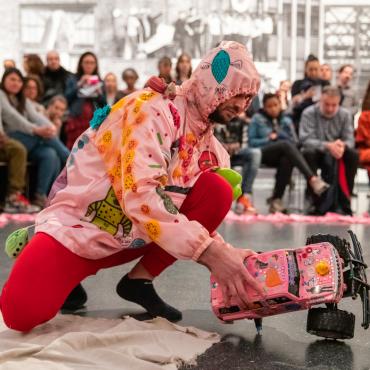 Christopher is squatting in the middle of a space outlined by pink cloth. He wears a pink hoodie and a red union suit and holds a broken remote control car. Both the car and the hoodie are decorated with different objects. Behind, the attentive audience watches the performance. Photograph taken at the Brooklyn Museum.