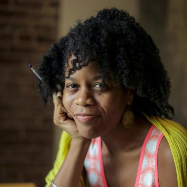 Ama Codjoe, a thirty-something Black woman poet, softly smiling at the camera, with a pen in her hand