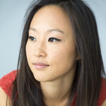 Crystal, a Korean-American woman in her thirties, is looking off to her left with a neutral expression on her face. Her long dark brown hair covers her shoulders. She is leaning towards the camera in a red-orange v-neck dress. Parts of her bare arms are visible.