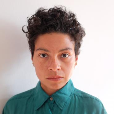 Mariana Valencia has brown skin and short black curly hair, she is framed from the chest up wearing a green shirt with buttons and collar; against a white wall.