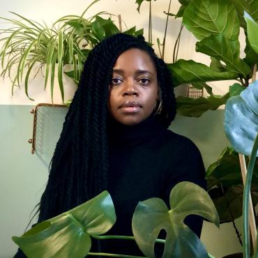 Diane Exavier center-framed wearing a black turtleneck surrounded by various houseplants