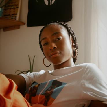 Sadé sitting on a chair looking into the camera wearing a t-shirt and orange skirt.