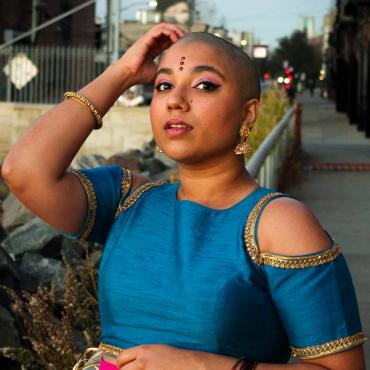 Bald femme person wearing a blue Indian crop top and a hot pink skirt looks into the camera with their hand on their head. They are wearing Indian jewelry and three pottus/bindis on their forehead.