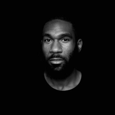 In a black and white portrait, Justin pools in a pitch black background. He's a twenty-something Black man with a full beard and a calm and intent gaze.