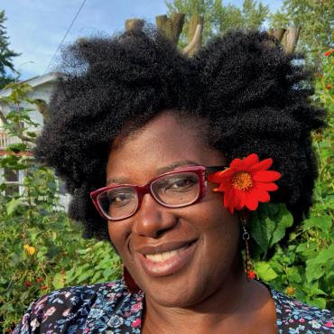 Kashimana Ahua a thirty-something has a flower in her afro hair and is smiling behind glasses in front of green trees.
