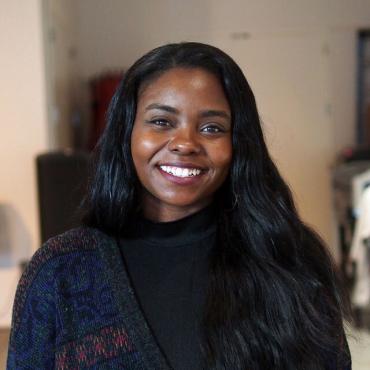 LaJuné McMillian, A 28-year-old, Black, and gender non conforming artist, smiles at the camera in Black Turtle neck, and dark blue cardigan.