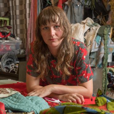 A picture of Lindsay Rhyner surrounded by fabric in her studio. She is holding a pair of scissors.
