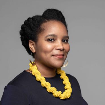 A bright smiling African femme with a yellow necklace, navy shirt, and gold hoop earrings. Grey background. This is a traditional headshot.