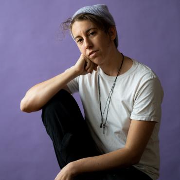 A white, genderqueer person wearing a white shirt and black pants leans their face on their hand pensively, sitting in front of a lavender backdrop.