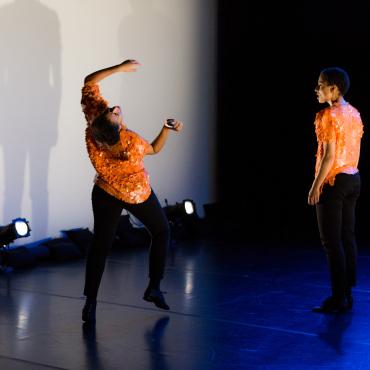 Twin brown women onstage wearing orange sequined tops. Cast in blue light, the twin to the left twists back with arms up, while her sister stands still in observation. Their shadows are doubled on a white side wall.