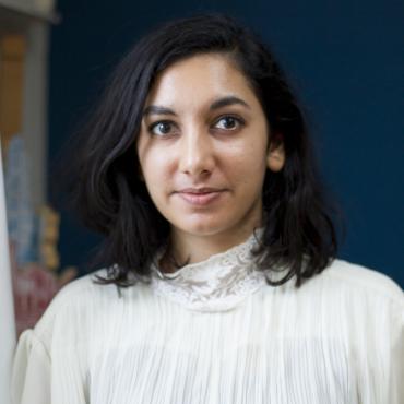 Sunita, a brown femme in a white blouse standing in front of a blue wall.