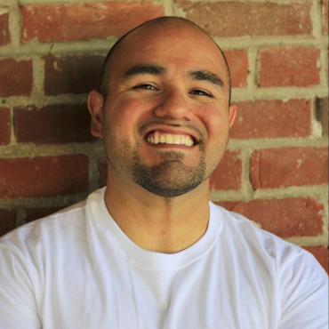 Michael Torres, a thirty-four-year old Chicano poet and prose writer stands in front of a brick wall with his arms crossed, smiling at the camera.