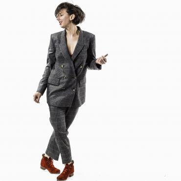 A white woman with brown short hair and red lipstick moving, feet crossed and head tilted. In a gray checked suit with red tap dance boots.