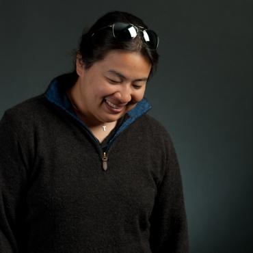 Images shows Valerie Oliveiro in a dark brown sweater, sunglasses on their head. They are looking downwards, quiet but playful. The background is black.