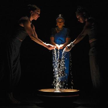 Three women pouring glass pebbles into a lit cauldron.