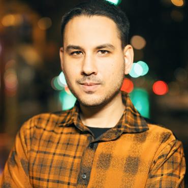 Suneil Sanzgiri, a light-skinned Indian American male, staring into the camera on a brightly lit street corner at night.