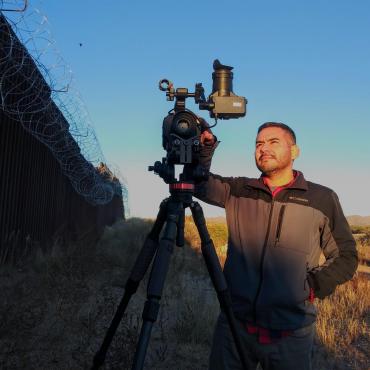 Rafael, a Latino man in his early 40's, looking through the camera lens on a tripod in front of the US/MX border. A bird is seen flying over the barb-wired border fence.