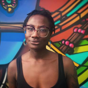 Raven Johnson, a thirty-one year old, Black woman with glasses standing in front of a colorful wall mural.