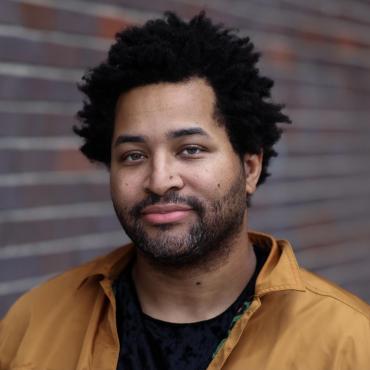 Headshot of Tommy Franklin, 37 year old Black man with black afro-ish hair, wearing a light brown jacket and black shirt.