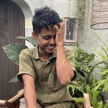 The artist (a brown trans person with short black hair, wearing a green linen shirt and light khaki pants) seated on an oak chair without a back against a background of gravel and plants.