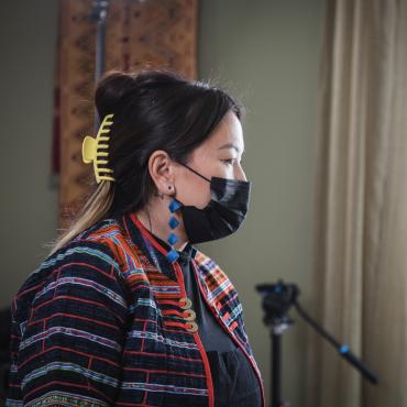 Profile image of a HMong person facing a film set. They are wearing a face mask with contemporary HMong earrings and upcycled jacket.