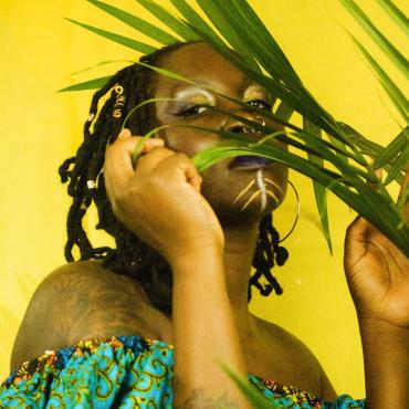 Atim Opoka, Early thirties dark skin black woman. With shoulder length locs. partly covered face by Green plant. in front of a yellow background.