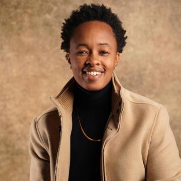 Tish Jones seated before a golden backdrop in a blue turtleneck and tan coat, smiling.