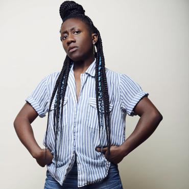nicHi douglas, a 30-something Black femme, stands powerfully with hands on hips in a blue striped shirt and well-worn denim.