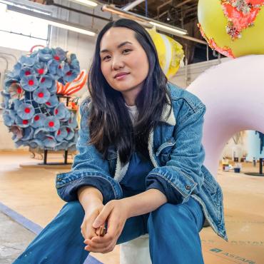 Amanda Phingbodhipakkiya, a thirty-something Thai and Indonesian woman artist in a warehouse looking at the camera flanked by her work.