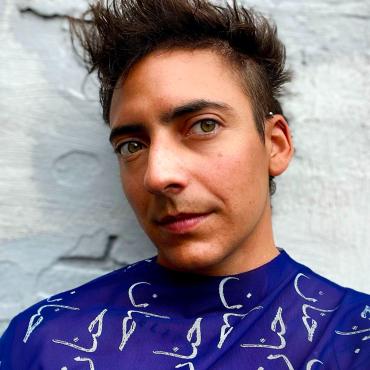 Headshot of a trans arab-american with spiky brown hair, wearing a long sleeve sapphire mesh with the word tender in arabic patterned in white.