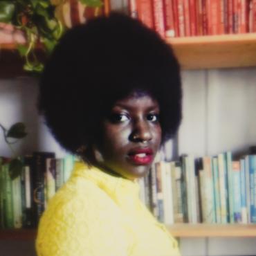 Angel Nafis, a thirty-something Black woman poet standing in front of a full bookshelf.