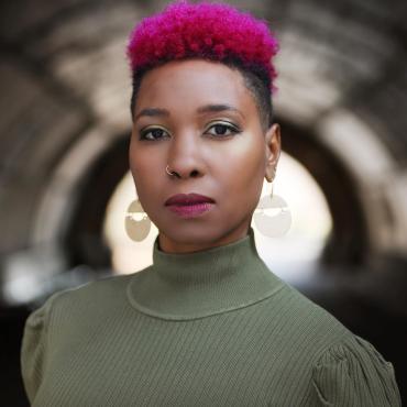 Kendra J. Ross, a thirty-something Black woman poses in front of a blurry tunnel. She has a pink afro hawk, gold earrings, and burgundy lipstick. She is wearing an olive green turtleneck sweater.