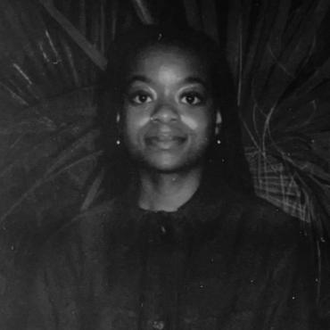 Crystal Kayiza, 29-year-old Black women film director, outside, standing in front of large plant, looking into camera.