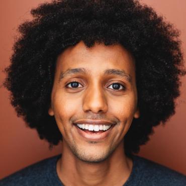 Sam Kebede, a young fresh faced black man with a large black afro, smiles to camera in a headshot with a black henley shirt in an auburn-colored headshot space.