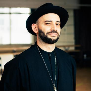 Christopher, a mixed-race man with pale skin, brown eyes, and a salt-and-pepper beard, looks straight into the camera. He wears a black suit and hat and silver jewelry. Behind him, a mirrored dance studio.